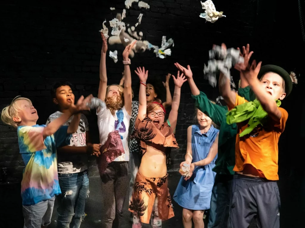 Flera glada barn från teaterskolan Konfetti står på scenen och kastar konfetti upp i luften.

Useat iloiset lapset teatterikoulu Konfetista seisoo lavalla ja heittävät konfettia ilmaan.