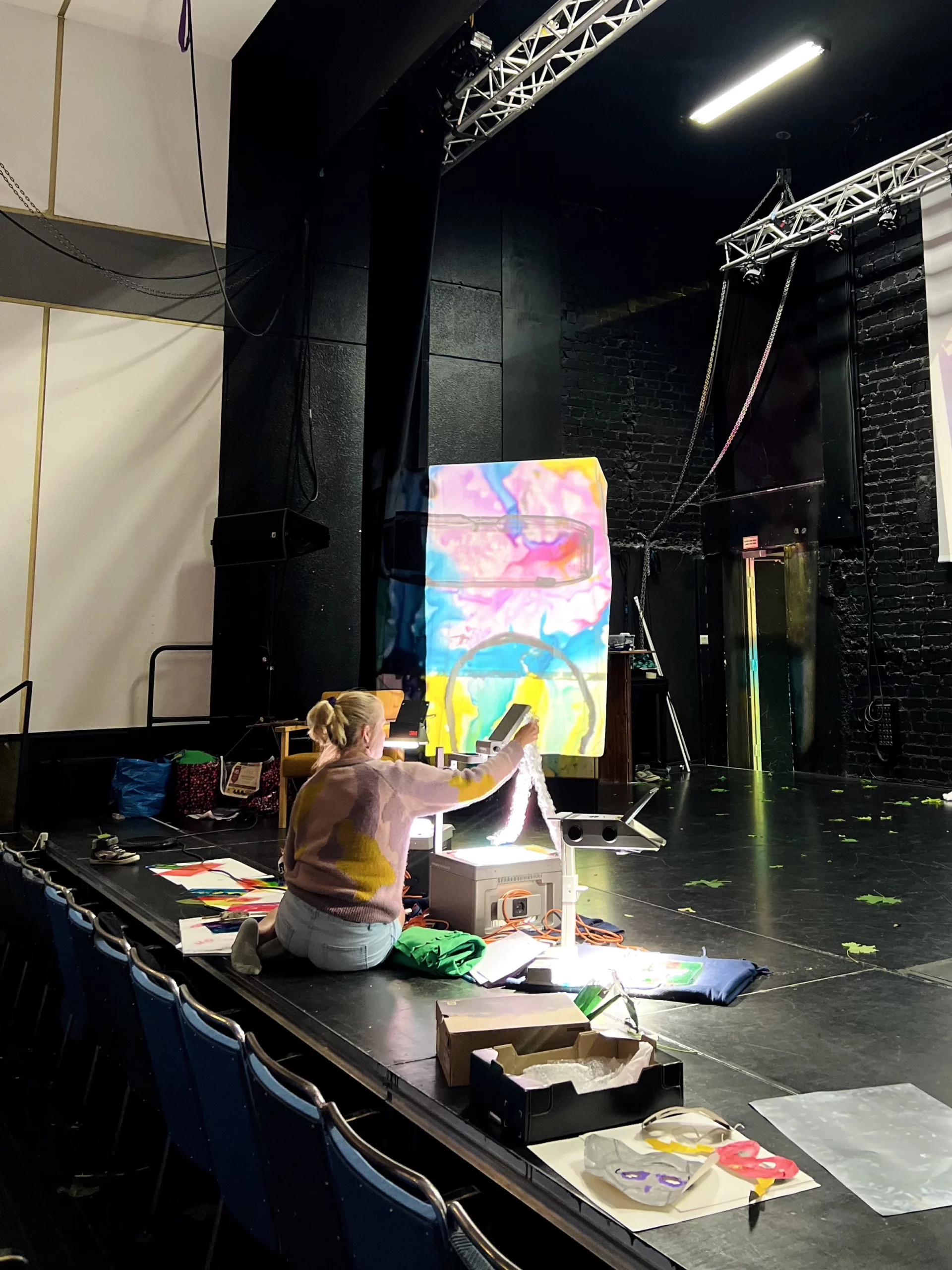 En konstnär sitter på Karelias scen och visar upp ett färgglatt konstverk med hjälp av en OH-apparat.

Taitelija istuu Karelian näyttämöllä ja esittää värikästä taidetta piirtoheittimen avulla.